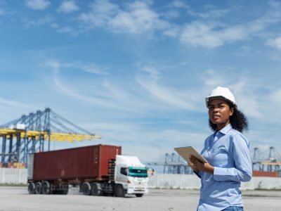 A female logistics officer in a hard hat observes cargo operations at a shipping port. Holding a tablet, she monitors container trucks and cranes involved in global transport and export logistics.